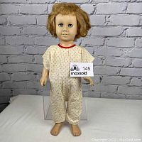 Front view of vintage Chatty Cathy doll standing on a white surface against a white brick background, showing outfit and face details.