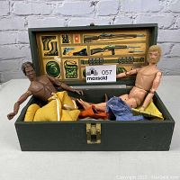 Two nude vintage GI Joe dolls seated inside a green wooden box with assorted clothing inside and illustrated military gear schematic on the lid.