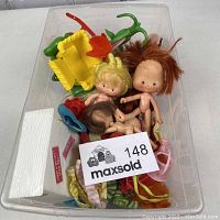 Top view of the clear plastic container holding three nude vintage Strawberry Shortcake dolls with hair in red, blonde, and brown colors surrounded by various doll accessories including yellow cart and pink watering can.