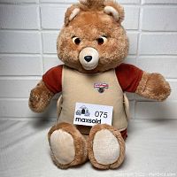 Full front view of vintage Teddy Ruxpin bear sitting against white brick wall, showing red shirt and beige vest with Teddy Ruxpin logo patch on chest.