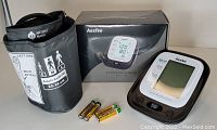 Photo of Aesfee electronic blood pressure monitor, two upper arm cuffs sized large and medium, and four AA batteries, all displayed on a flat surface with packaging box in background.