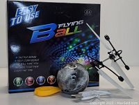 Photo showing the B Flying Ball toy drone, its packaging box, remote control, charging cable, and wrist strap arranged together.