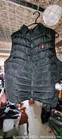 Small black Genovega heated vest hanging on a rack showing front side with quilting and red heat control button on chest.