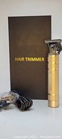 Photo showing a gold metal hair trimmer with a professional T blade head, accompanied by a charging cord and black box labeled 'HAIR TRIMMER'.