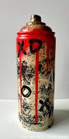 Front side of the spray can featuring red background on top with black 'X', 'O', 'X' symbols with red paint drips over a textured beige and black speckled surface.