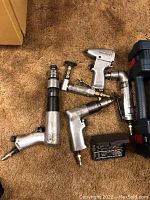Five silver pneumatic air tools on carpet: long straight air drill, pistol-grip air drill, inline die grinders, right-angle drill, right-angle grinder with green buffing pad.