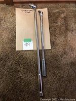 Two chrome breaker bars of different lengths laid on floor with handwritten label "Breaker Bars". Longer bar has fixed head and shorter has swivel head.