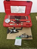 Open red plastic case showing pressure gauge, flexible hoses, and metal adapters organized inside with instruction label on lid.