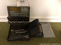 Photo showing three drill bit sets: one large metal case, a smaller black plastic case with orange interior, and a small clear plastic case containing various drill bits.