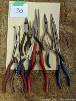 Photo showing 9 various needle nose and other specialized pliers with assorted handle colors and designs laid flat on beige surface
