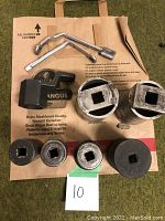 Photo of miscellaneous large sockets and tools arranged on paper bag, showing curved lever tool, ratchet extension bar, and various sizes of sockets with square openings.