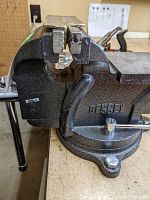 Close-up photo of gray cast metal bench vice mounted on a workbench, showing serrated jaws and textured finish.