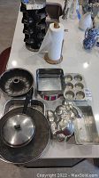 Overview of bakeware and cookware items arranged on table including bunt cake pan, cupcake pan, bunny cake pan, square baking pan, loaf pan, frying pan with lid, and more