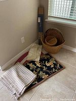 Photo showing three area rugs folded and stacked on the floor, multiple woven baskets nearby, and an unopened boxed drapery rod leaning against the wall