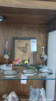 Glass shelf holding two metal bird figurines, one ceramic bird figurine, textured metal bird plaque, and three round glass ashtrays