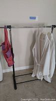 Plastic clothing rack with two white robes hanging and a cloth multi-pocket beach bag attached to the bar.