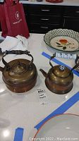 Two copper teapots shown on a kitchen counter with blue tape near them; larger teapot has wooden handle and smaller teapot has white and blue ceramic handle and knob.