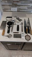 Collection of kitchen utensils spread on countertop with power outlet and tile backsplash in background, showing assortment of cutting boards, utensils and tools.