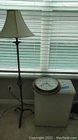 Floor lamp beside white wicker hamper with large round clock resting on top, photographed in front of a window with blinds.