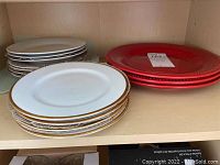 Multiple stacks of plates and saucers on a shelf, including 7 white stoneware dinner plates, 4 red stoneware dinner plates, 3 white saucers with gold rims, and 3 plain white saucers. Items show signs of use and need cleaning.