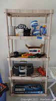 Front view of the 5-tier plastic shelving unit with various items on shelves for size reference. Shelving is beige plastic with tubular supports and slotted shelves.