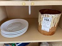 Four white ceramic plates stacked with a detailed rabbit motif embossed on each and an Italian pottery vase with a brown drip glaze beside them, placed on a wooden shelf.