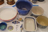 Photo showing various bowls including a large blue bowl, white bowl with orange rim, striped bowl, floral patterned bowl, and ceramic fish wall decor pieces on table.