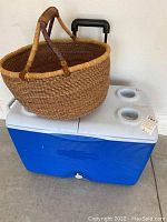 Photo showing the blue and white Rubbermaid cooler with two built-in cup holders on the lid and a wicker basket resting on top.