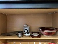 Shelf with sugar bowl with cat face lid, small metal creamer, wooden trivet, coaster, and red ceramic bowl.