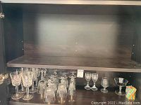 Shelf view showing nearly 20 different clear glass stemware pieces arranged in rows, including wine glasses, smaller stemmed glasses, and coupe glasses