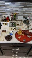 Countertop with multiple items including basket with pinecones and napkin rings, ceramic trivets, wooden tray with teapots, figurines and small decorative pieces.