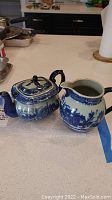Blue and white ironstone teapot and creamer set displayed on a countertop, showing scenic building transferware pattern with dark blue handles