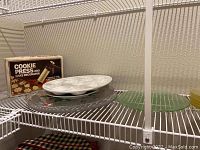 Shelf with boxed cookie press and cake decorator, white ceramic plate, clear oval glass tray, green round glass tray