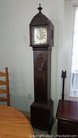 Full length view of tall antique grandmother clock showing solid mahogany case, pedestal base, detailed upper door panel with carved decoration, finial on top, and clock face enclosed behind glass.