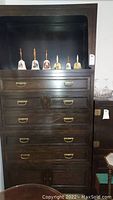 Front view of fully assembled dark wood tall cabinet with drawers and open shelf at top, displaying decorative collectible bells on the shelf.