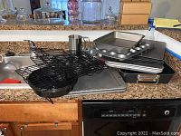 Various baking and cooking pans, cooling racks, flour sifter, and roasting pan arranged on kitchen counter.