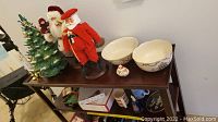 Full view of ceramic lighted Christmas tree, Santa figurines, Lenox bowls and mugs on wooden shelf.