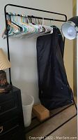 Metal clothes rack with assorted hangers, black garment bag hanging from it, white trash can and small box underneath