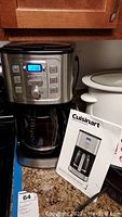 Photo of Cuisinart coffee maker CBC-7000PC on counter showing front panel with digital display and buttons, glass carafe, and instruction manual.
