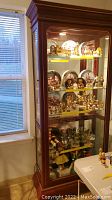 Front and left side view of the wooden curio cabinet showing glass shelves with various collectibles inside and light at the top.