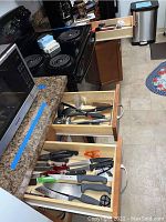 Two kitchen drawers opened showing various kitchen knives and cutters, some with black and red handles, placed in wooden drawer dividers on a granite countertop.