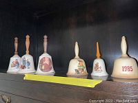 Front view of six collectible ceramic bells on a shelf with different designs and handles including wood and ceramic.