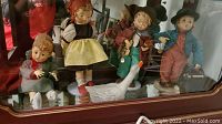 Four dolls arranged standing and seated along with a duck figurine inside a display cabinet.