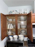 Open kitchen cabinet showing stacks of plates, bowls, mugs and an oval platter on top of the cabinet