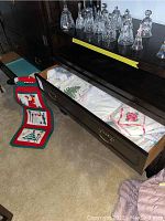 Open drawer with folded white linens including tablecloths and pillowcases, and festive Christmas card holder on floor.