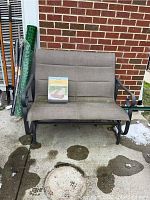 Front view of outdoor porch glider bench with beige fabric and metal frame, showing some shadowing and dirt on ground.