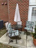 Patio set on concrete slab against brick wall and window. Shows table with closed umbrella and four chairs, two with cushions.