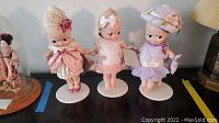Full view of 3 Kewpie dolls on white stands, dressed in pastel colors with accessories