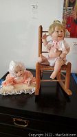 Two porcelain dolls side by side, one sitting in a wooden rocker and the other lying on a pillow