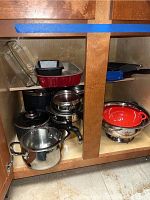 Open cabinet showing multiple pots, pans, glass baking dishes stacked inside and red metal colanders on the right side.